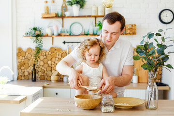 A small child girl is playing in the bright Scandinavian-style kitchen at the table. Father and daughter play with grits. The concept of Montessori education. Eco kitchen.