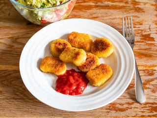 portion of fried Chicken nuggets from frozen product (deep-fried breaded chicken meat) with tomato ketchup on white plate and salad from fresh vegetables on old wooden table in home kitchen