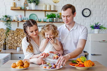 Family portrait of a happy mother, daughter and father posing in the kitchen during Breakfast, eating delicious macaroons, cakes, cookies. Married good relations, love each other
