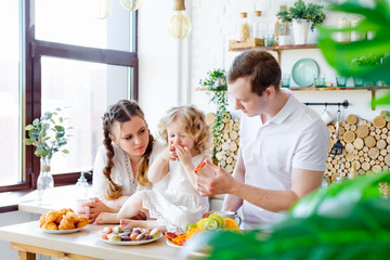 Family portrait of a happy mother, daughter and father posing in the kitchen during Breakfast, eating delicious macaroons, cakes, cookies. Married good relations, love each other