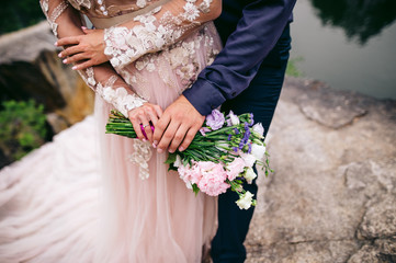 Bride and groom are standing on the rock near the water hugging, bride holds wedding flowers in her...