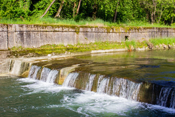Obraz premium Spring landscape. The threshold of a mountain meandering river enclosed in stone fences. The water drops rapidly. Green plants, shrubs, and trees. Sunny weather.