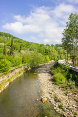 Fototapeta premium Spring landscape. A mountain meandering river enclosed in stone fences. Shallow, with a bare stone bottom on the side. Green plants, shrubs, and trees. Sunny weather.