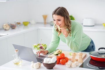 Portrait of her she nice attractive lovely pretty girl eating avocado cooking vegs using laptop watching tutorial video quarantine self isolation tasting flavor in modern light white kitchen house