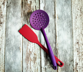 Kitchen utensils. Red plastic kitchen spatula and violet plastic slotted spoon on background with aged wooden boards. - Powered by Adobe