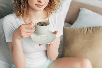cropped view of young woman in homewear holding cup of coffee on bed in morning