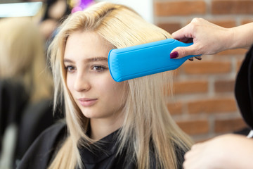 Portrait of a beautiful blonde girl in the interior of a beauty salon. The master hairdresser makes styling with a curling iron after successful dyeing of straight hair. Beautiful woman is smiling.