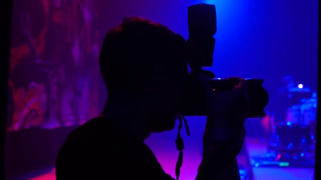 A young male photographer holds a camera backstage taking pictures on a stage with a blue and red background light in the dark. Reportage shooting of the concert.