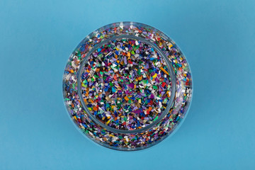 Crushed plastic pellets particles made of multicolored recycled plastics collected at sea in a glass jar, blue background ready to be used for new circular products. Waste particles, reduce pollution