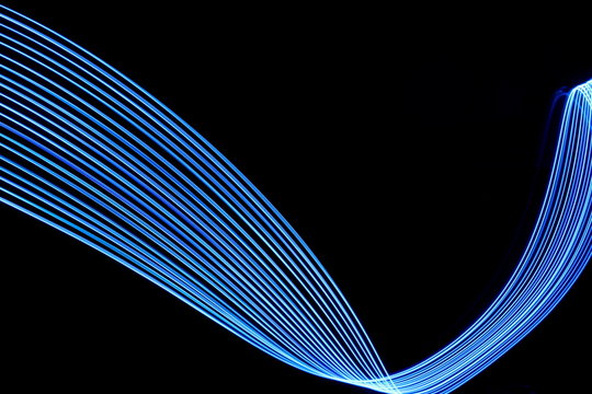 Long exposure photograph of neon electric blue colour in an abstract swirl, parallel lines pattern against a black background. Light painting photography.