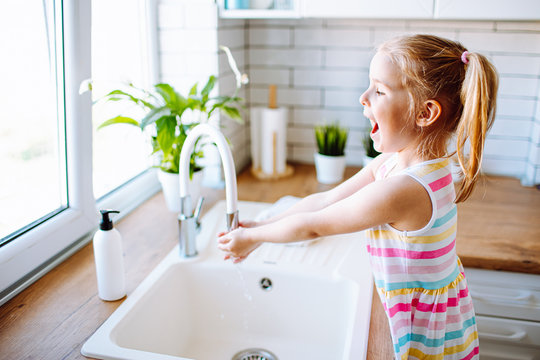 Blonde toddler girl washing hands in the light kitchen before eating. Hygiene and healthcare concept.