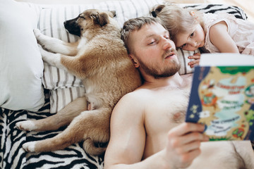 A man of European appearance reads a book for a night a girl of three years. The dog is sleeping on the bed. Father and daughter.