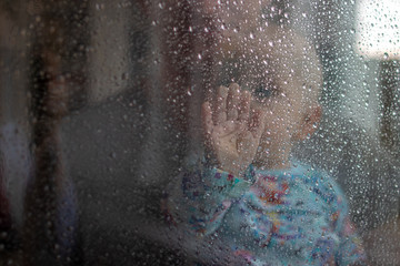 White baby boy looking through glass window on the rain drops and rain outside and pressing his small palm against the glass - isolation, quarantine, virus outside, stay safe, stay home (palm covering