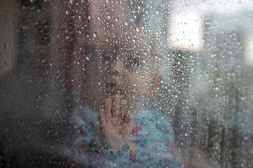 White baby boy looking through glass window on the rain drops and rain outside and pressing his small palm against the glass - isolation, quarantine, virus outside, stay safe, stay home - portrait