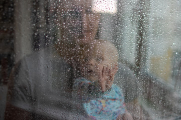 White baby boy looking through glass window on the rain drops and rain outside and pressing his small palm against the glass - isolation, quarantine, virus outside, stay safe, stay home (from distance