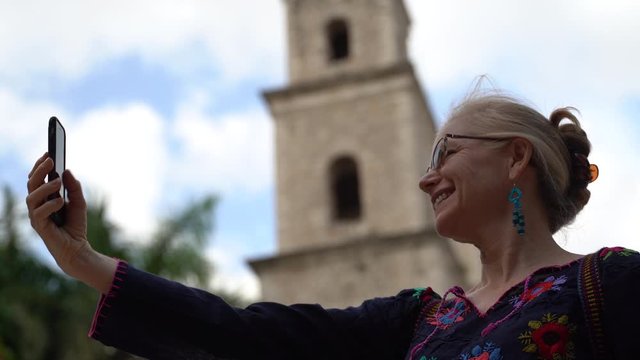 Holding out smartphone, a beautiful blond middle aged mature woman smiles and takes a selfie in Hidalgo park in Merida, Museum.
