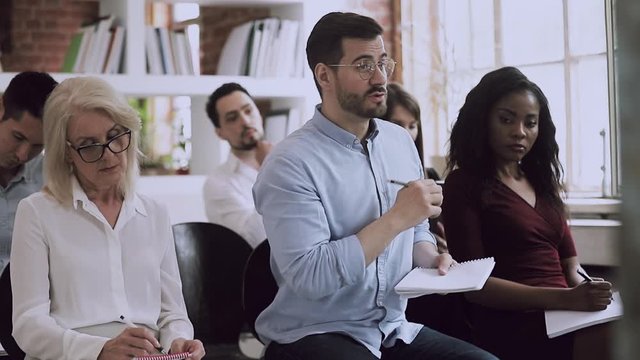 Confident young businessman in eyeglasses taking part in educational lecture with different ages mixed race people, raising hand, asking question to speaker during corporate workshop at office.
