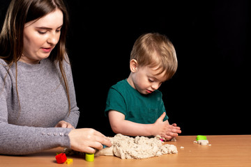 Little child with mom play kinetic sand on the table. multi-colored shapes nearby. hands closeup. home family leisure