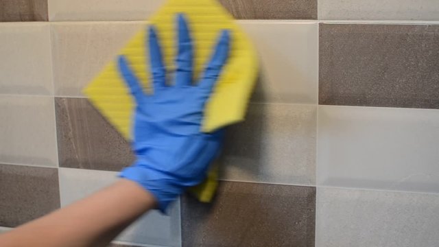 A housemaid in rubber gloves is washing a ceramic tile on a wall with a rag and cleaning agent