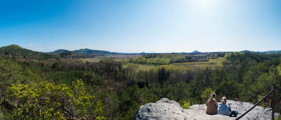two woman sitting at sandstone rock viewpoint Havrani skaly, spring landscape in Lusatian Mountains...