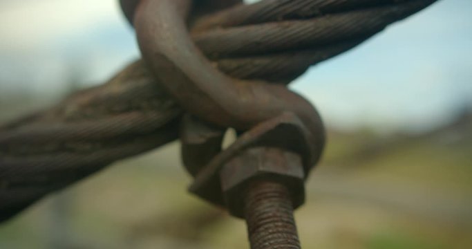 Old rusty coarse thread bolt with huge hooked nut secures woven steel cable of suspension pedestrian bridge against blurred background of grass and sky. Close-up The wire rope divides frame diagonally