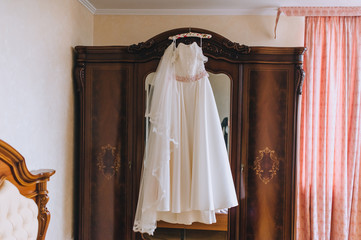 Wedding white long dress of the bride hangs on a hanger, on a wooden wardrobe with a mirror in the hotel room. Photography, concept.