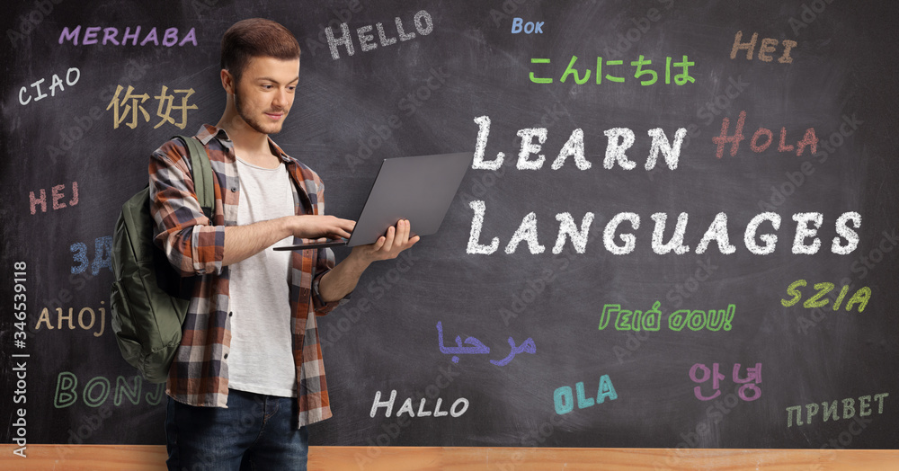 Wall mural male student wiht a laptop and standing in front of a blackboard with text learn languages