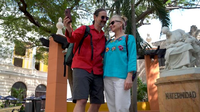 Mature, middle aged couple smiles for a selfie photo using smartphone in front of the Maternity sculpture in Hidalgo park in Merida, Mexico.