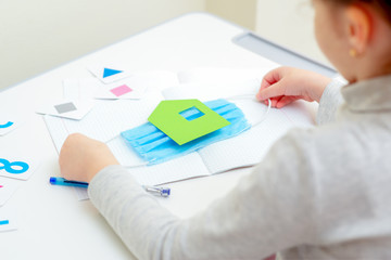 Hands of child holding a medical mask with paper green house over copybook during studying at home. Home learning concept.