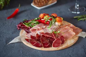 Antipasto on a wooden plate close-up. Cold smoked meat plate with sliced ham, prosciutto, and bacon. Decorated with bruschetta with fresh vegetables and herbs