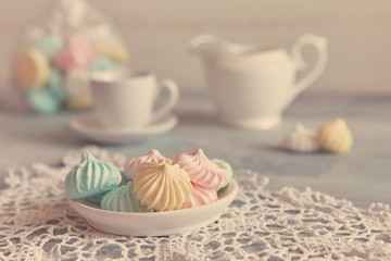 Multi-colored meringues in pastel shades with cups and a jug on a light textured wooden background. The concept of Breakfast with tea or coffee, home comfort and tenderness. Close-up, selective focus