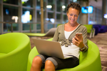 Obraz premium Happy mature beautiful tourist woman using phone and laptop at the lounge indoors