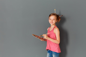 A cute little girl with a palette and brushes against a gray wall is going to write a greeting for mother's day
