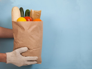 Male hands in cotton gloves hold a paper bag with pasta, tomatoes, cucumbers, daikon, lemon, orange on a blue background. Zero waste. Courier delivery. Volunteer.