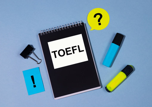 TOEFL English test exam written in black notebook with clips, markers and memo notes around on blue table