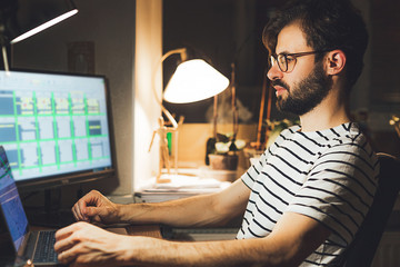 Modern young man working remotely from home at night. Casual millennial guy typing on pc notebook surfing internet looking at screen enjoying distant job