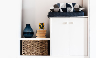 Cozy home interior decor: pillow, plaid, blue vase, stack of books, cup of coffee, wicker box on a white shelf in the room. The quarantine concept of stay home, distance home education background.