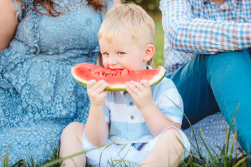 The baby has a watermelon in nature. A little boy with white hair sits on grass with his parents. Picnic.