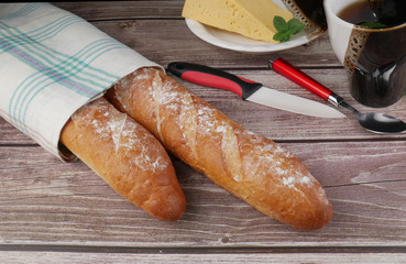 Two bread baguettes wrapped in a white towel on a wooden table. In the background is a plate with...