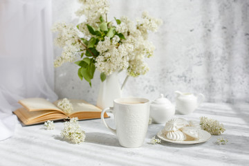 Fototapeta na wymiar Still life vase with a bouquet of white lilac, a Cup of coffee with marshmallows, a plate of meringue, a sugar bowl and a milk jug , an old book. Postcard good morning.