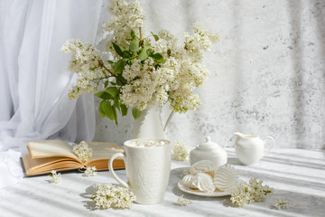 Still life vase with a bouquet of white lilac, a Cup of coffee with marshmallows, a plate of meringue, a sugar bowl and a milk jug , an old book. Postcard good morning.