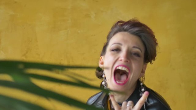 Let's rock concept. Portrait of excited beautiful woman with dark pink hair in casual yellow dress standing, looking at camera, showing rock and roll gesture.