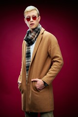 Handsome young man in stylish sunglasses and scarf posing against red studio background 