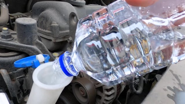 Man pours water or special cleaning solution from plastic bottle into a car window washer tank, and closes the lid, close up. Independent unprofessional automobile maintenance during a road trip.