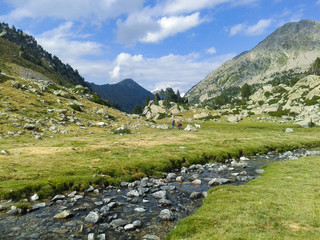 Two people hiking in the middle of a high mountain valley with a small river surrounded by trees with the mountains and sky behind. Horizontal image. © Marx Bamala