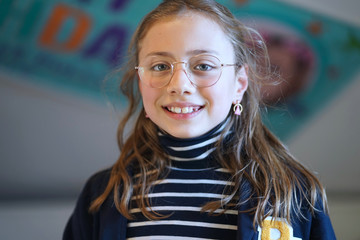 Portrait of a 10 year old schoolgirl with blue white striped turtle neck jersey, college jacket, glasses and blonde hair smiling at the camera.   