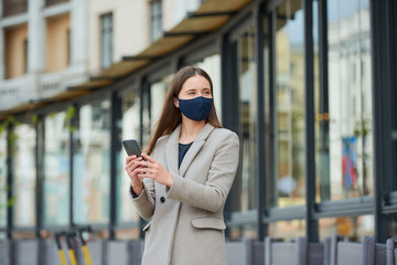 A girl with long hair in a navy blue face mask to avoid the spread coronavirus uses a smartphone in the street. A woman in a face mask against COVID-19 wears a coat looks away in the city.