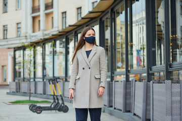 A brunette girl with long hair in a navy blue face mask to avoid the spread coronavirus holds a smartphone in the street. A woman in a face mask against COVID-19 wears a coat looks away in the city.