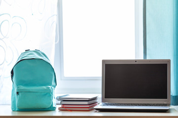 Blue school backpack with school supplies and computer on table in children room across from the window in sunny day.