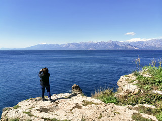 A woman in a dark blue jacket stands on a high rocky shore above the sea against the backdrop of the mountains and looks into the distance. Image with selective focus.